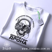 Футболка оператор дрона білого кольору, зображена зверху, на ній підвішений етикет, фон світлий, принт з черепом у навушниках з окулярами, текст "ОПЕРАТОР ДРОНА" під ним.