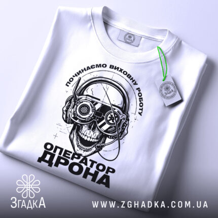 Футболка оператор дрона білого кольору, зображена зверху, на ній підвішений етикет, фон світлий, принт з черепом у навушниках з окулярами, текст "ОПЕРАТОР ДРОНА" під ним.