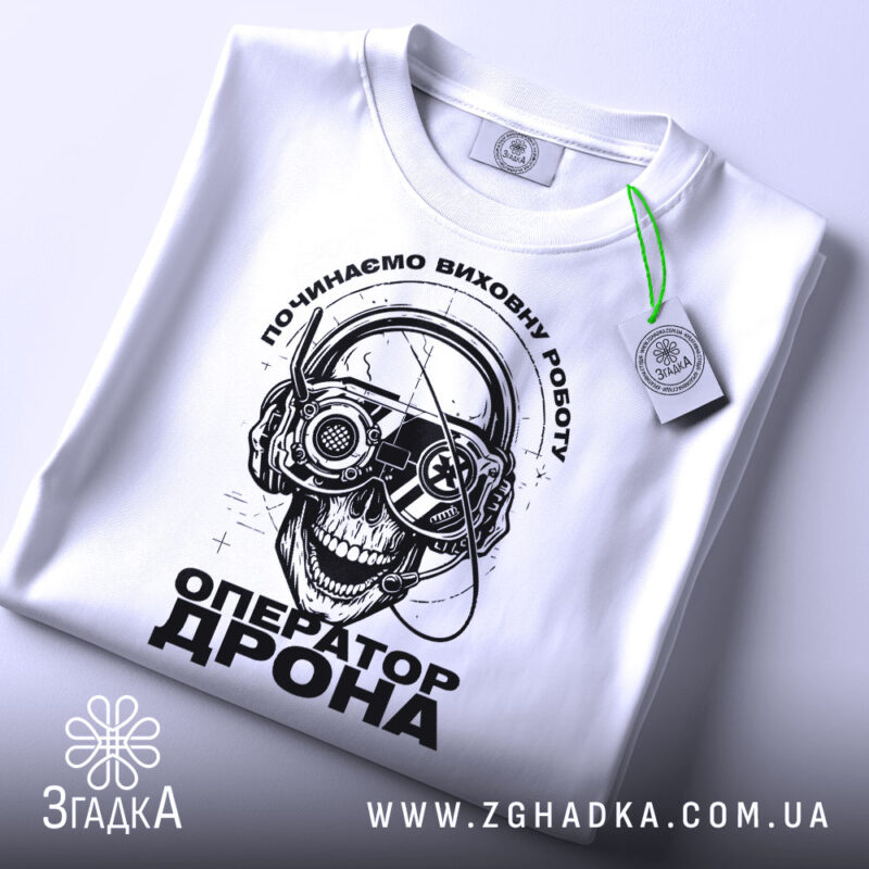Футболка оператор дрона білого кольору, зображена зверху, на ній підвішений етикет, фон світлий, принт з черепом у навушниках з окулярами, текст "ОПЕРАТОР ДРОНА" під ним.