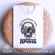 Футболка оператор дрона білого кольору, зображена зверху на плетеній основі, фон світлий, принт з черепом у навушниках з окулярами, текст "ОПЕРАТОР ДРОНА" під ним.