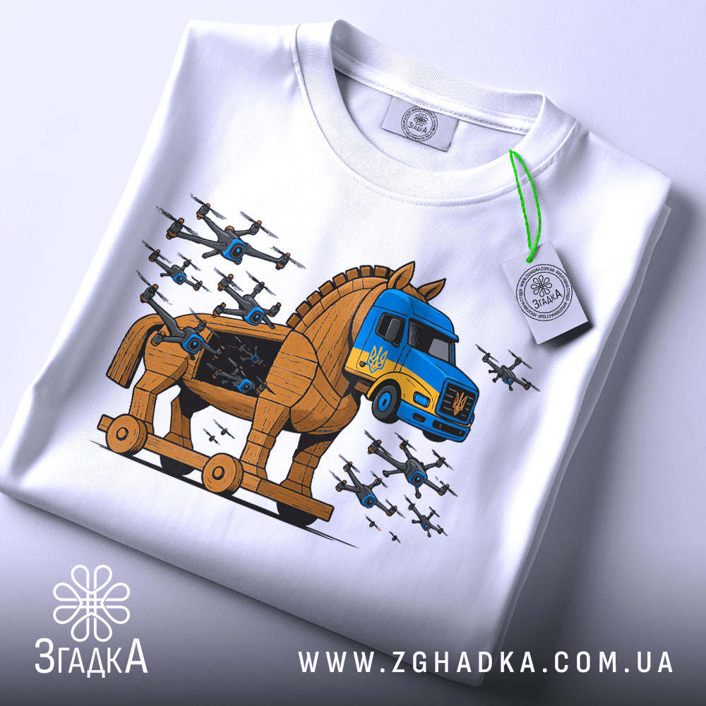 Футболка операція павутина подарунок. Біла футболка, складена на столі, з принтом, що зображує Троянського коня та вантажівку. На футболці видно етикетку з зеленим шнурком. Фон світло-сірий, деталі принта яскраві, з літаками.