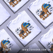 Футболка операція павутина подарунок. Біла футболка на манекені, з принтом, що зображує дерев'яного коня Троянського, який перевозить вантажівку з українським гербом. Фон світло-сірий, деталі принта яскраві, з літаками, що оточують коня.