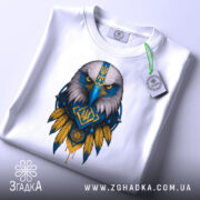 Футболка орел Україна, біла, зображена зверху, фон світло-сірий, принт з орлом в синьо-жовтих кольорах, на комірці видно етикетку з логотипом.