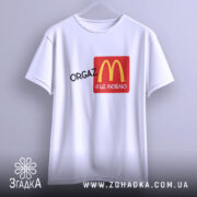 Футболка оргазм я це люблю, біла футболка підвішена на вішалці, з принтом логотипу McDonald's у червоному квадраті, текст 'ORGAZ' чорного кольору, фон світло-сірий.