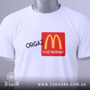 Футболка оргазм я це люблю, складена біла футболка з принтом, на якому зображено логотип McDonald's у червоному квадраті, текст 'ORGAZ' чорного кольору, на футболці є етикетка з зеленим шнурком, фон світлий.
