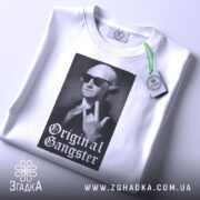 Футболка оригінальний ґангстер білого кольору, складена на поверхні, з видимим етикеткою, фон світло-сірий, принт з зображенням чоловіка в окулярах, який показує жест, з написом 'Original Gangster' під ним.