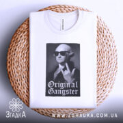 Футболка оригінальний ґангстер білого кольору, складена на плетеній кошику, фон світло-сірий, принт з зображенням чоловіка в окулярах, який показує жест, з написом 'Original Gangster' під ним.