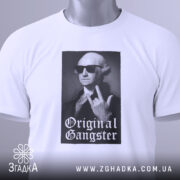 Футболка оригінальний ґангстер білого кольору, складена на поверхні, з видимим етикеткою, фон світло-сірий, принт з зображенням чоловіка в окулярах, який показує жест, з написом 'Original Gangster' під ним.
