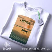 Футболка озеро Світязь, біла, складена на поверхні, фон світло-сірий, принт з малюнком озера, човна та сонця, текст на принті зеленого кольору, з етикеткою з зеленим шнурком.