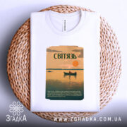 Футболка озеро Світязь, біла, складена на плетеній основі, фон світло-сірий, принт з малюнком озера, човна та сонця, текст на принті зеленого кольору.