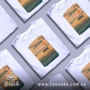 Футболка озеро Світязь, біла, зображена на манекені, фон світло-сірий, принт з малюнком озера, човна та сонця, текст на принті зеленого кольору.