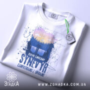 Футболка озеро Синевир білого кольору, зображена зверху, на ній видна етикетка з зеленим шнурком, фон світлий, принт зображає ліс та озеро з написом 'Озеро Синевир SYNEVYR Carpathians / Ukraine'.