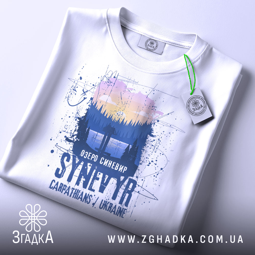 Футболка озеро Синевир білого кольору, зображена зверху, на ній видна етикетка з зеленим шнурком, фон світлий, принт зображає ліс та озеро з написом 'Озеро Синевир SYNEVYR Carpathians / Ukraine'.
