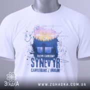 Футболка озеро Синевир білого кольору, зображена зверху, на ній видна етикетка з зеленим шнурком, фон світлий, принт зображає ліс та озеро з написом 'Озеро Синевир SYNEVYR Carpathians / Ukraine'.