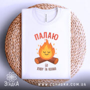 Футболка палаю кохаю, біла, зображена зверху на плетеному кошику, на фоні світло-сірого градієнту, принт з вогнем та колодами, текст червоним шрифтом.