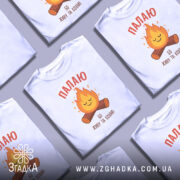 Футболка палаю кохаю, біла, зображена з фронтального ракурсу, на фоні світло-сірого градієнту, принт з веселим вогнем та дерев'яними колодами, текст червоним шрифтом.