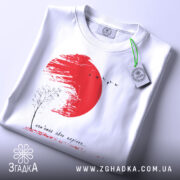 Футболка памʼятай своє коріння, білого кольору, складена на поверхні, фон світлий, принт з червоним сонцем, чорним деревом та птахами, прикріплений ярлик з логотипом.