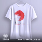 Футболка памʼятай своє коріння, білого кольору, підвішена на вішалці, фон світлий, принт з червоним сонцем, чорним деревом та птахами, текст під малюнком.