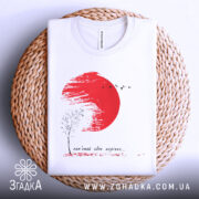 Футболка памʼятай своє коріння, білого кольору, складена на плетеній кошику, фон світлий, принт містить червоне сонце, чорне дерево та птахів, текст під малюнком.