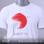 Футболка памʼятай своє коріння, білого кольору, складена на поверхні, фон світлий, принт з червоним сонцем, чорним деревом та птахами, прикріплений ярлик з логотипом.