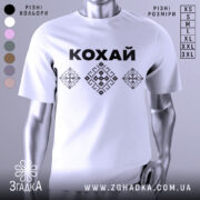 Футболка патріотична кохай, білого кольору, зображена на манекені, фон нейтральний, принт з чорною написом 'КОХАЙ' та вишиванковими елементами у вигляді ромбів.