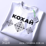 Футболка патріотична кохай, білого кольору, складена на столі, фон світлий, принт з чорною написом 'КОХАЙ' та вишиванковими елементами, на комірці видно етикетку.