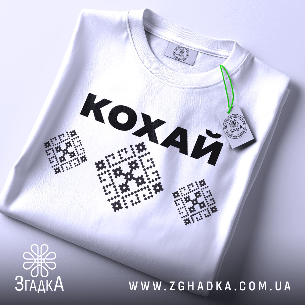 Футболка патріотична кохай, білого кольору, складена на столі, фон світлий, принт з чорною написом 'КОХАЙ' та вишиванковими елементами, на комірці видно етикетку.