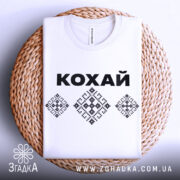 Футболка патріотична кохай, білого кольору, складена на плетеній кошику, фон світлий, принт з чорною написом 'КОХАЙ' та вишиванковими елементами, видна етикетка на комірці.
