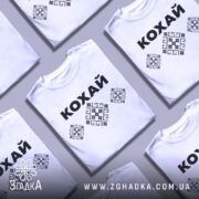 Футболка патріотична кохай, білого кольору, зображена на манекені, фон нейтральний, принт з чорною написом 'КОХАЙ' та вишиванковими елементами у вигляді ромбів.