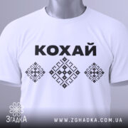 Футболка патріотична кохай, білого кольору, складена на столі, фон світлий, принт з чорною написом 'КОХАЙ' та вишиванковими елементами, на комірці видно етикетку.