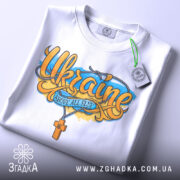 Футболка патріотична Україна, біла, складена на плоскій поверхні, фон світло-сірий, принт з написом 'Ukraine' та жовто-блакитними елементами, на ярлику зелена стрічка.