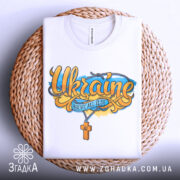 Футболка патріотична Україна, біла, складена на плоскій поверхні, фон світло-сірий, принт з написом 'Ukraine' та жовто-блакитними елементами, розташована на плетеній основі.