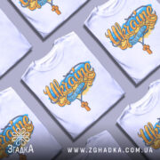 Футболка патріотична Україна, біла, зображена на манекені з правого боку, фон світло-сірий, принт містить напис 'Ukraine' з жовто-блакитними елементами та хрестом.