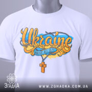 Футболка патріотична Україна, біла, складена на плоскій поверхні, фон світло-сірий, принт з написом 'Ukraine' та жовто-блакитними елементами, на ярлику зелена стрічка.