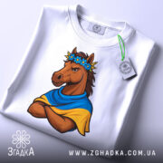 Футболка патріотичний кінь, біла, складена на поверхні, фон світло-сірий, принт з кінем у вінку з квітів та синьо-жовтому шарфі, кінь з серйозним виразом обличчя, на футболці є етикетка.