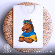 Футболка патріотичний кінь, біла, складена на кошику, фон світло-сірий, принт з кінем у вінку з квітів та синьо-жовтому шарфі, кінь з серйозним виразом обличчя, видно етикетку на горловині.