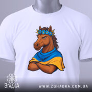 Футболка патріотичний кінь, біла, складена на поверхні, фон світло-сірий, принт з кінем у вінку з квітів та синьо-жовтому шарфі, кінь з серйозним виразом обличчя, на футболці є етикетка.