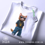 Футболка патріотичний кіт, біла, зображена з верхнього ракурсу, на фоні світло-сірого градієнту, принт з котом у темній футболці з жовтим гербом України та синіх штанах, в темних окулярах, з етикеткою на зеленому шнурку.