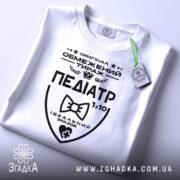 Футболка педіатр білого кольору, складена на плоскій поверхні, фон світло-сірий, принт чорного кольору з написом 'ПЕДІАТР' та етикеткою з зеленим шнурком.