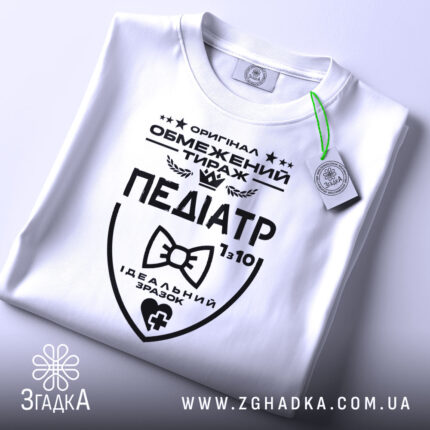 Футболка педіатр білого кольору, складена на плоскій поверхні, фон світло-сірий, принт чорного кольору з написом 'ПЕДІАТР' та етикеткою з зеленим шнурком.