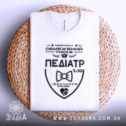 Футболка педіатр білого кольору, складена на плоскій поверхні з плетеного матеріалу, фон світло-сірий, принт чорного кольору з написом 'ПЕДІАТР' та графічними елементами.