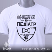 Футболка педіатр білого кольору, складена на плоскій поверхні, фон світло-сірий, принт чорного кольору з написом 'ПЕДІАТР' та етикеткою з зеленим шнурком.