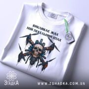 Футболка пекельний птах, біла, складена на поверхні, фон світло-сірий, принт з черепом та пропелерами, на футболці є етикетка з зеленим шнурком.