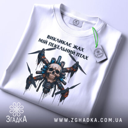 Футболка пекельний птах, біла, складена на поверхні, фон світло-сірий, принт з черепом та пропелерами, на футболці є етикетка з зеленим шнурком.