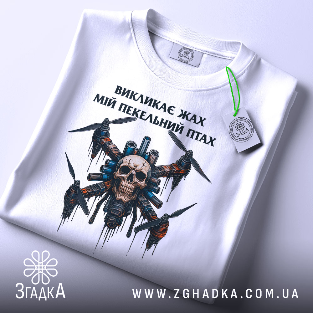 Футболка пекельний птах, біла, складена на поверхні, фон світло-сірий, принт з черепом та пропелерами, на футболці є етикетка з зеленим шнурком.