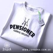 Футболка пенсіонер кайфую, біла, зображена зверху, на фоні світло-сірого кольору, з принтом чорного кольору, що містить слово 'PENSIONER' та малюнок візка, з етикеткою зеленого кольору.
