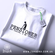Футболка пенсіонер ще рухаюсь, біла, складена на поверхні, з верхнього ракурсу, фон світло-сірий, принт чорного кольору з написом 'PENSIONER' та малюнком людини з палицею, на ярлику зелена стрічка.