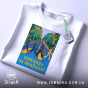 Футболка переходь на українську, біла, зображена в горизонтальному ракурсі, фон світло-сірий, принт з п'ятьма персонажами, що переходять дорогу, на фоні зелених дерев, текст "ПЕРЕХОДЬ НА УКРАЇНСЬКУ" у жовтому кольорі, на комірці є етикетка з логотипом.