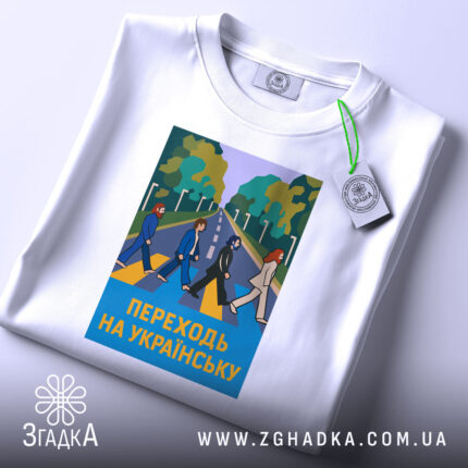 Футболка переходь на українську, біла, зображена в горизонтальному ракурсі, фон світло-сірий, принт з п'ятьма персонажами, що переходять дорогу, на фоні зелених дерев, текст "ПЕРЕХОДЬ НА УКРАЇНСЬКУ" у жовтому кольорі, на комірці є етикетка з логотипом.