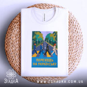 Футболка переходь на українську, біла, зображена на плетеній кошику, фон світло-сірий, принт з п'ятьма персонажами, що переходять дорогу, на фоні зелених дерев, текст "ПЕРЕХОДЬ НА УКРАЇНСЬКУ" у жовтому кольорі, комірець видимий.